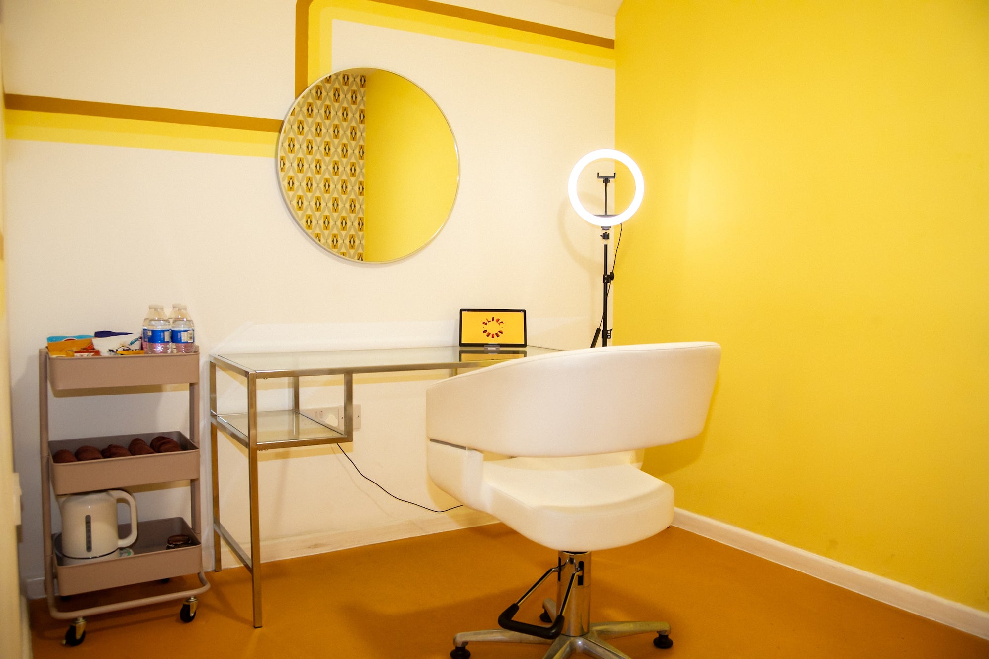 Studios beauty pod with yellow wall, a desk, chair, ring light, snack tray and mirror