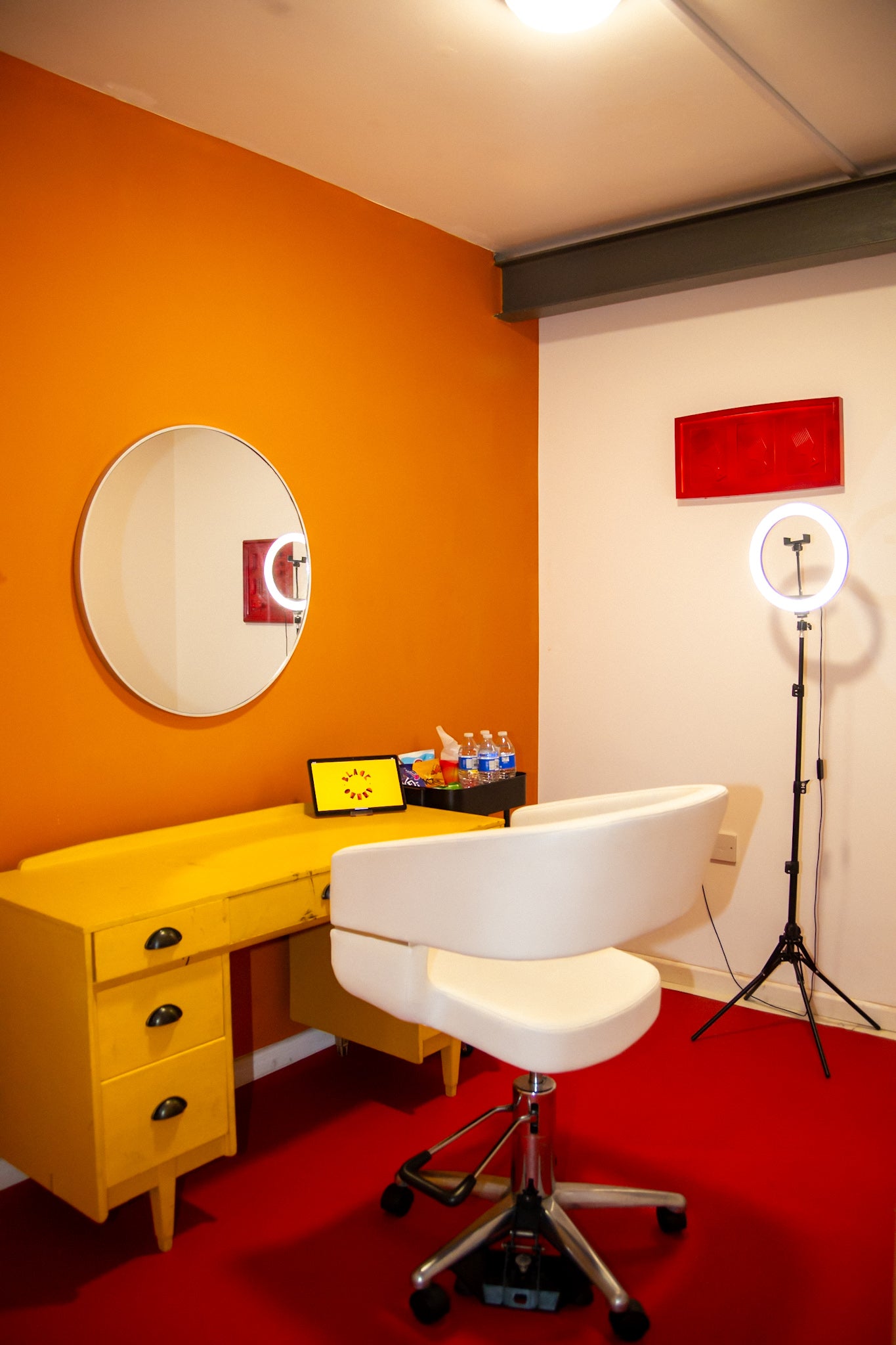 Studios space with yellow desk, white chair in a room with orange walls, a red floor, ring light and a mirror.